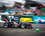 Ein Rennwagen mit der Nummer 70 auf der Seite, auf der stolz das Logo der Hochschule Coburg zu sehen ist, rast an verschwommenen Zuschauern und Bannern vorbei. Der Fahrer trägt einen Helm, das Auto hat einen großen Heckflügel und ein elegantes schwarz-rotes Design. Die Szene vermittelt ein aufregendes Gefühl von Bewegung und Aufregung.