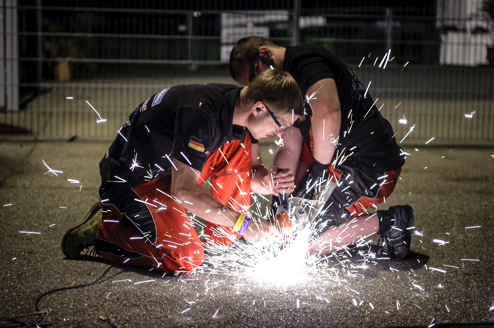 Zwei Menschen kauern auf dem Boden, tragen Schutzbrillen und arbeiten mit Metall, während Funken um sie herumfliegen. Es ist Nacht, beleuchtet von einer hellen Schweißlampe auf einem Außengelände der Hochschule Coburg, hinter ihnen ein Zaun.
