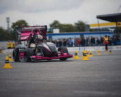 Ein Rennwagen mit rosa und schwarzen Akzenten, der stolz die Hochschule Coburg repräsentiert, manövriert durch einen mit gelben Pylonen markierten Hindernisparcours. Der Fahrer trägt einen Helm. Im Hintergrund stehen Zuschauer bei einem Baldachin und verfolgen gespannt die spannende Darbietung.