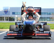 Eine Person in Rennanzug und Helm steht in einem geparkten Formel-Rennwagen auf dem Campus der Hochschule Coburg und hält einen Fußball über dem Kopf. Auf der Heckklappe des Autos sitzt eine Plüschkatze, im Hintergrund ragt ein großes Gebäude auf.