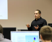 Ein Mann im schwarzen Hemd hält an der Hochschule Coburg einen Vortrag. Er hält ein Gerät in der Hand und gestikuliert, während mehrere Leute vor ihm sitzen und ihre Augen auf den Bildschirm gerichtet halten, auf dem eine bunte Grafik angezeigt wird.