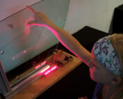 Ein Kind mit einem Stirnband interagiert mit einem Wissenschaftsexponat der Hochschule Coburg, indem es eine Glasplatte berührt, die mit rotem Licht beleuchtet wird. Der Aufbau besteht aus elektronischen Komponenten und einem Holzsockel und suggeriert eine pädagogische Ausstellung oder ein interaktives Experiment.