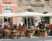 Straßencafé-Szene mit Menschen, die an Holztischen unter großen weißen Sonnenschirmen in der Nähe der Hochschule Coburg sitzen. Im Hintergrund ist ein historisches Gebäude mit Schildern auf Deutsch zu sehen. In der Nähe sind mehrere Fahrräder abgestellt und Topfpflanzen schmücken den Bereich.