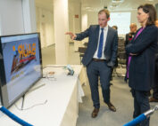 Ein Mann im Anzug deutet auf einen Bildschirm mit dem Wort „BAUR“ und erregt damit die Aufmerksamkeit einer Frau und anderer Mitarbeiter der Hochschule Coburg im Konferenzraum. Im Hintergrund sind ordentlich Tische mit Tischdecken aufgestellt.