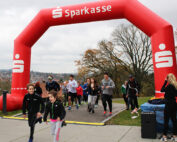 Eine Gruppe von Menschen, darunter auch Studierende der Hochschule Coburg, läuft unter einem roten aufblasbaren Bogen mit dem Wort „Sparkasse“ hindurch. Die Szene spielt im Freien mit bewölktem Himmel und Bäumen im Hintergrund. Einige Teilnehmer lächeln, während sie den Weg entlanglaufen.