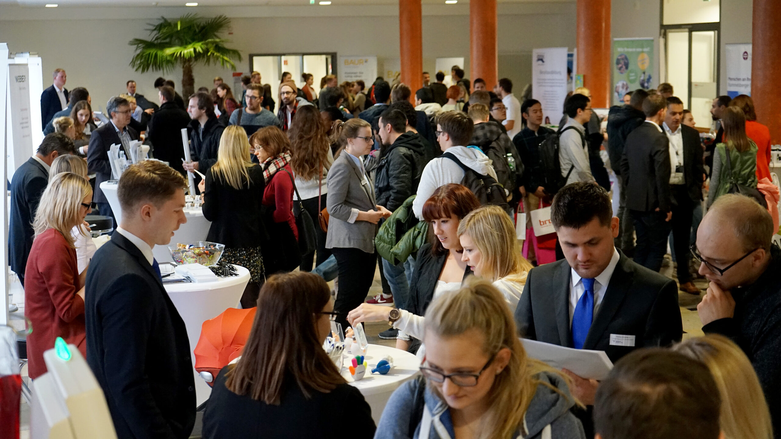 Auf der geschäftigen Karrieremesse der Hochschule Coburg herrscht ein pulsierendes Treiben, während Teilnehmer in Business-Kleidung Gespräche führen und sich inmitten der lebhaften Atmosphäre im Innenbereich vernetzen. An den Tischen liegen Broschüren und Werbeartikel bereit, um Kontakte in diesem lebhaften akademischen Zentrum zu knüpfen.