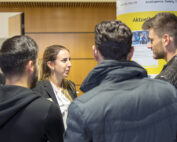 Bei der Veranstaltung der Hochschule Coburg unterhält sich eine Frau im Business-Anzug mit vier Männern, die der Kamera den Rücken zukehren. Sie stehen vor einem Display mit teilweise lesbarem Text, was eine Konferenzatmosphäre in Innenräumen suggeriert.