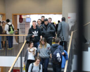 Eine Gruppe von Menschen, darunter Männer und Frauen, geht in einem belebten Innenbereich der Hochschule Coburg eine Treppe auf und ab. Einige tragen Rucksäcke und legere Kleidung wie Jacken und Sweatshirts. Der Bereich ähnelt einem öffentlichen Raum, möglicherweise innerhalb der lebhaften Campusumgebung.