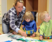 Ein Erwachsener und zwei kleine Kinder sind an der Hochschule Coburg bei einer Malaktivität dabei. Die Kinder sitzen in Schutzkitteln mit Pinseln auf dem Boden, während der Erwachsene ihnen hilft, ihre künstlerischen Ausdrucksformen auf einem großen weißen Blatt zu lenken. Farbenfrohe Kunst schmückt den Hintergrund.