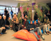 Eine Gruppe von Menschen posiert in Innenräumen der Hochschule Coburg. Einige sitzen auf bunten Sitzsäcken, während andere knien oder herumstehen. Sie befinden sich in einer entspannten Umgebung mit einer großen Palme im Hintergrund und großen Fenstern an der Seite. Alle wirken entspannt und lächeln.