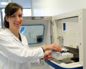 Eine Person im Laborkittel bedient eine wissenschaftliche Maschine und lächelt, während sie im Advanced Laboratory der Hochschule Coburg, umgeben von verschiedenen hochmodernen Geräten, ein Tablett einschiebt.