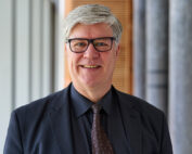 Ein Mann mit kurzen grauen Haaren und Brille, vermutlich ein Fakultätsmitglied der Hochschule Coburg, lächelt in die Kamera. Er trägt einen dunklen Anzug, ein schwarzes Hemd und eine gemusterte Krawatte. Im Hintergrund ist ein modernes Interieur mit Säulen und vertikalen Jalousien zu sehen, das ein akademisches Ambiente widerspiegelt.