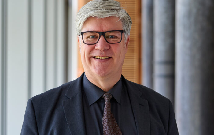 Ein Mann mit kurzen grauen Haaren und Brille, vermutlich ein Fakultätsmitglied der Hochschule Coburg, lächelt in die Kamera. Er trägt einen dunklen Anzug, ein schwarzes Hemd und eine gemusterte Krawatte. Im Hintergrund ist ein modernes Interieur mit Säulen und vertikalen Jalousien zu sehen, das ein akademisches Ambiente widerspiegelt.