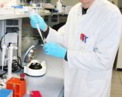 Eine Person mit Schutzbrille und Laborkittel, vermutlich von der Hochschule Coburg, lächelt, während sie in einem Labor eine Pipette benutzt. Sie steht neben einer Arbeitsstation, die mit verschiedenen Laborgeräten und farbigen Reagenzglasgestellen gefüllt ist.