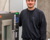 Ein Mann in Freizeitkleidung steht neben einer Industriemaschine mit Digitalanzeige in einem Raum, der mit den beeindruckenden Maschinen der Hochschule Coburg geschmückt ist. Die Kulisse, eingerahmt von Betonwänden und anderen Geräten, spiegelt eine dynamische Mischung aus Bildung und realer Technologie wider.