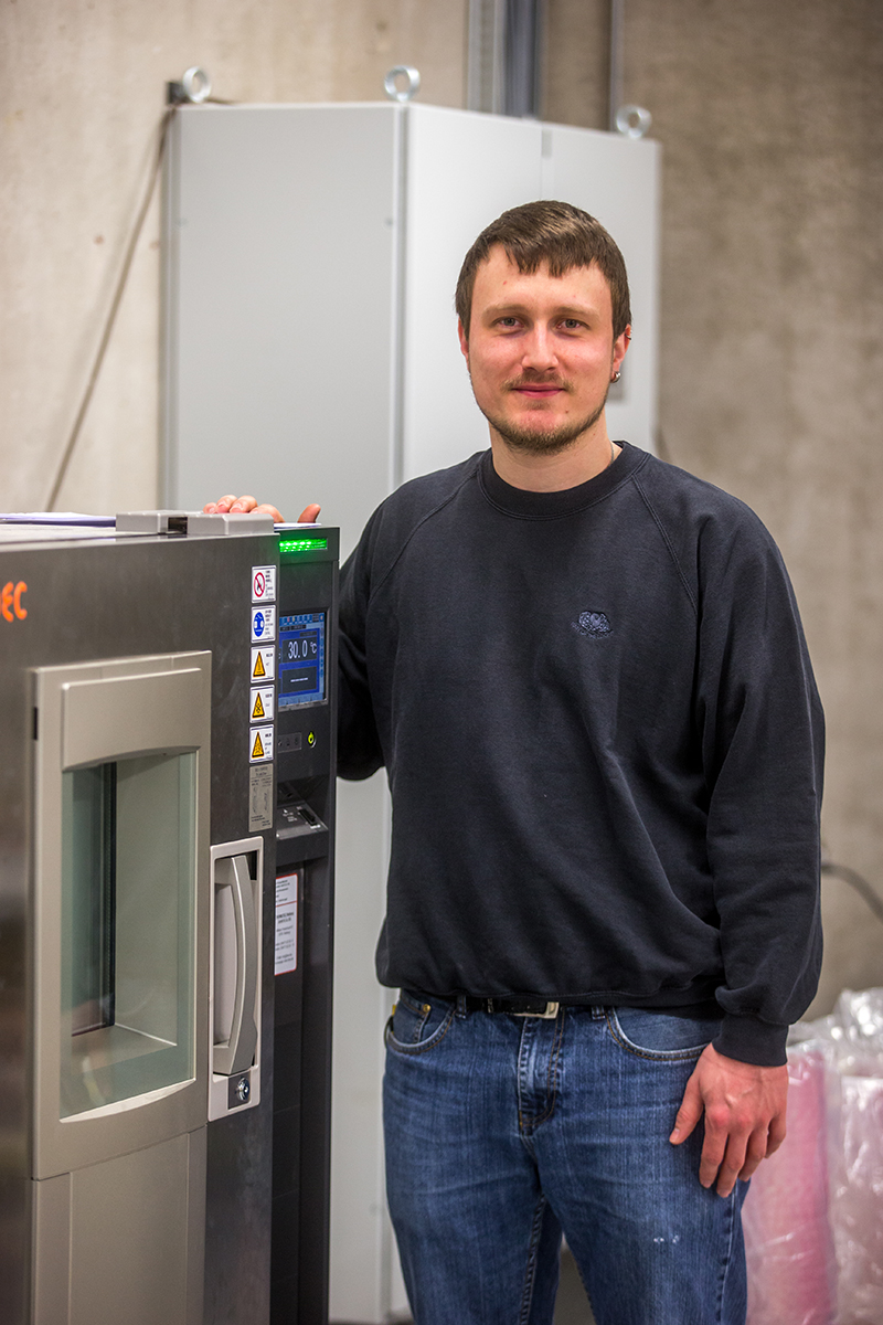 Ein Mann in Freizeitkleidung steht neben einer Industriemaschine mit Digitalanzeige in einem Raum, der mit den beeindruckenden Maschinen der Hochschule Coburg geschmückt ist. Die Kulisse, eingerahmt von Betonwänden und anderen Geräten, spiegelt eine dynamische Mischung aus Bildung und realer Technologie wider.