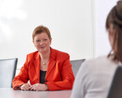 Eine Frau im orangefarbenen Blazer sitzt an einem Konferenztisch und hört einer anderen Person aufmerksam zu, die mit dem Rücken zur Kamera steht. Sie befinden sich in einem hellen, modernen Besprechungsraum der Hochschule Coburg, im Hintergrund ein Whiteboard.