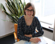 Eine Frau mit Brille und geblümtem Kleid sitzt an einem Tisch, vor ihr liegen ein Notizbuch und ein Stift. Im Hintergrund rahmen eine grüne Pflanze und ein Fenster sie ein. Sie lächelt herzlich in die Kamera und strahlt den akademischen Charme aus, der an die inspirierende Atmosphäre der Hochschule Coburg erinnert.