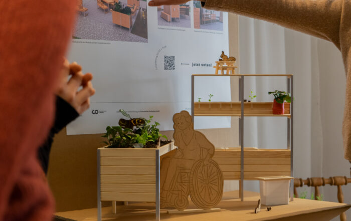 Eine Person deutet auf ein kleines Modell eines barrierefreien Gartens an der Hochschule Coburg. Das Modell besteht aus einem erhöhten Pflanzkasten aus Holz mit einer Figur im Rollstuhl, Regalen mit kleinen Pflanzen und einem Poster im Hintergrund, auf dem entsprechende Bilder zu sehen sind.