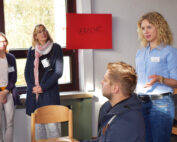 Eine Frau steht in einem Raum mit Stühlen und spricht mit einem sitzenden Mann, während zwei Frauen der Hochschule Coburg an einem Fenster stehen und zuschauen. An der Wand hinter ihnen hängt ein rotes Schild mit Text.