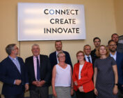 Vor einem Schild mit der Aufschrift „Connect Create Innovate“ steht eine Gruppe in Business-Kleidung, von denen einige lächeln und andere in die Kamera blicken. Sie repräsentiert den Geist der Hochschule Coburg. Die Inneneinrichtung vor einer beigen Wand unterstreicht ihr Engagement für akademische und professionelle Spitzenleistungen.