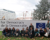 Eine Gruppe von Menschen der Hochschule Coburg sitzt und kniet im Gras vor einem großen Schild mit der Aufschrift „Weltforum für Demokratie, Europarat“. Gebäude und Herbstbäume bilden eine malerische Kulisse.
