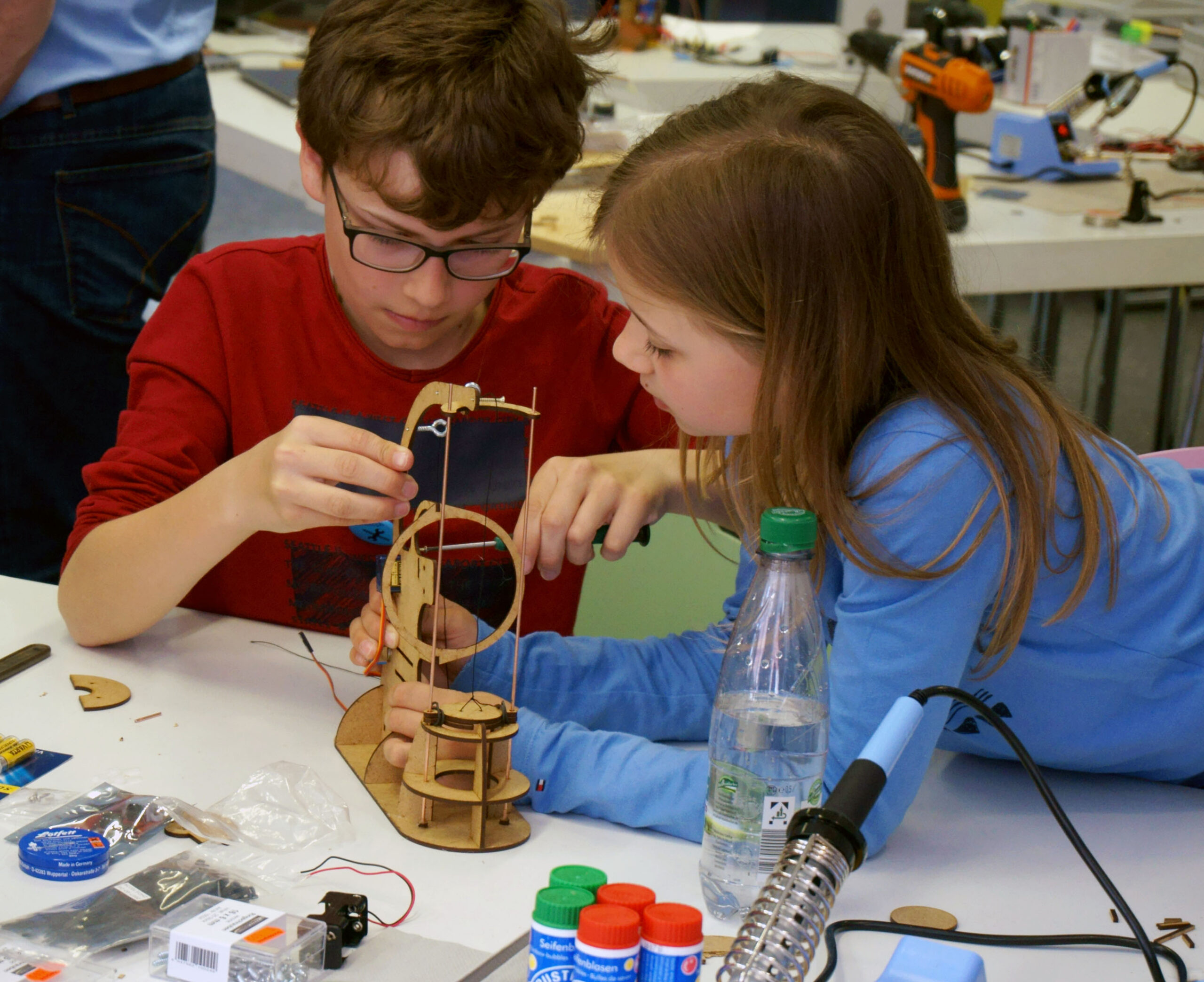 Zwei Kinder sind konzentriert dabei, an einem Tisch voller Bastelwerkzeuge und Materialien ein Holzmodell zusammenzusetzen, was an eine Werkstatt der Hochschule Coburg erinnert. Der Junge trägt eine Brille und ein rotes Hemd, während das Mädchen ein blaues Hemd trägt. Ein Lötkolben und andere Utensilien liegen verstreut um sie herum.