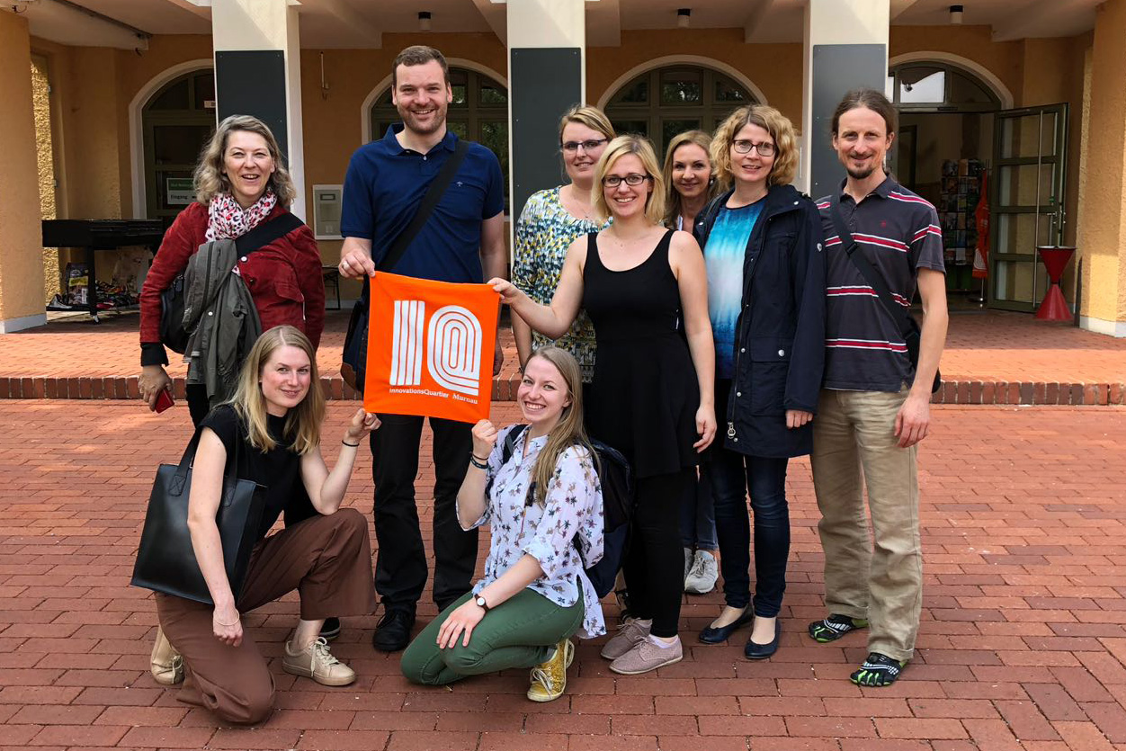 Eine Gruppe von acht Personen lächelt und posiert für ein Foto auf einem Ziegelweg vor einem Gebäude der Hochschule Coburg. Zwei Personen in der Mitte halten eine orangefarbene Tasche mit einem weißen Logo.