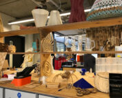 In einer Werkstatt der Hochschule Coburg sind auf Holzregalen verschiedene 3D-gedruckte und lasergeschnittene Modelle ausgestellt. Die Sammlung umfasst abstrakte Formen, Tiere und ein Schachbrett. Im Hintergrund schaffen Tische, Geräte und Werkzeuge eine inspirierende Atmosphäre für Innovationen.