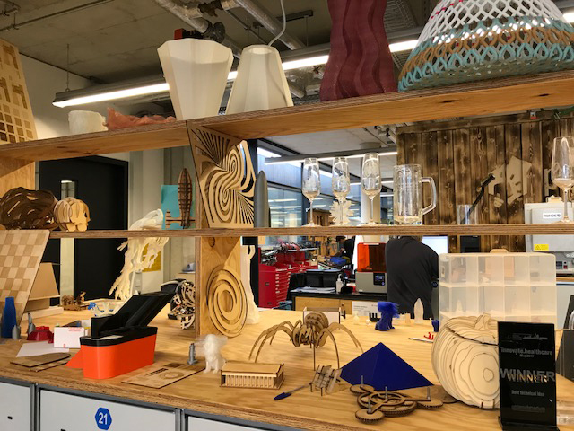 In einer Werkstatt der Hochschule Coburg sind auf Holzregalen verschiedene 3D-gedruckte und lasergeschnittene Modelle ausgestellt. Die Sammlung umfasst abstrakte Formen, Tiere und ein Schachbrett. Im Hintergrund schaffen Tische, Geräte und Werkzeuge eine inspirierende Atmosphäre für Innovationen.