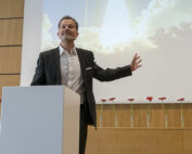 Ein Mann im schwarzen Anzug spricht an einem Rednerpult, im Hintergrund ist eine Leinwand mit Wolken und Wolkenkratzern zu sehen. Er wendet sich an das Publikum der Hochschule Coburg. Hinter ihm steht eine Reihe roter Blumen in Glasvasen auf einem Holzsims.
