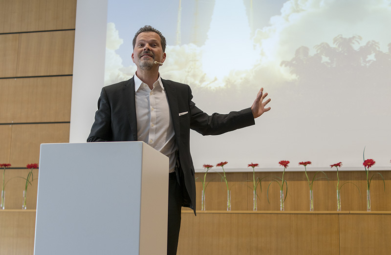 Ein Mann im schwarzen Anzug spricht an einem Rednerpult, im Hintergrund ist eine Leinwand mit Wolken und Wolkenkratzern zu sehen. Er wendet sich an das Publikum der Hochschule Coburg. Hinter ihm steht eine Reihe roter Blumen in Glasvasen auf einem Holzsims.