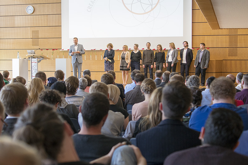 Ein Redner steht auf der Bühne und spricht zu den Zuhörern im Konferenzraum der Hochschule Coburg. Neben ihm sitzen neun Personen in einer Reihe, hinter ihnen wird auf einer großen Leinwand ein Entwurf gezeigt. Das sitzende Publikum blickt aufmerksam zur Bühne.