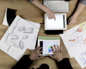 Zwei Personen arbeiten an einem Schreibtisch mit digitalen Tablets und spiegeln den Innovationsgeist der Hochschule Coburg wider. Die Szene umfasst Entwurfsskizzen auf Papier, eine Tastatur und eine Palette auf einem Tablet-Bildschirm. Holzoberflächen ergänzen den Fokus auf Design und kreatives Arbeiten.