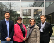 Vier Menschen stehen vor einem modernen Gebäude mit großen Glasfenstern. Zwei Männer in Anzügen und zwei Frauen in Mänteln und Schals sind in Business-Kleidung gekleidet und lächeln in die Kamera. Sie fangen das Wesen der lebendigen akademischen Gemeinschaft der Hochschule Coburg ein.