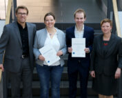 Vier Personen stehen auf einer Treppe im Innenbereich der Hochschule Coburg. Zwei in der Mitte halten lächelnd Zertifikate in den Händen, während zwei weitere auf beiden Seiten stehen. Sie sind in Business-Kleidung gekleidet und posieren für ein Gruppenfoto.