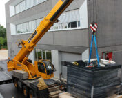 Vor dem modernen, mehrstöckigen Gebäude der Hochschule Coburg hebt ein großer gelber Kran eine Palette an, die mit schwarzem Material und blauen Gurten umwickelt ist. Eine Person steht daneben und leitet den Vorgang auf der gepflasterten Fläche.