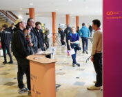 In einer geräumigen Halle versammelt sich eine Gruppe um einen Stand mit der Aufschrift „Hochschule Coburg“, während jemand mit ihnen spricht. In der Nähe schlendert eine Person mit Unterlagen vorbei, während andere im Hintergrund verkehren und zur lebhaften Atmosphäre beitragen.