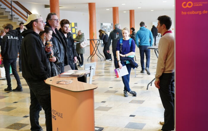 In einer geräumigen Halle versammelt sich eine Gruppe um einen Stand mit der Aufschrift „Hochschule Coburg“, während jemand mit ihnen spricht. In der Nähe schlendert eine Person mit Unterlagen vorbei, während andere im Hintergrund verkehren und zur lebhaften Atmosphäre beitragen.