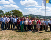 Eine bunt gemischte Gruppe von Menschen steht unter blauem Himmel auf einer Wiese im Freien zusammen und verkörpert den Geist der Hochschule Coburg. Sie posieren vor einer malerischen Kulisse mit Bäumen und einem Hügel in der Ferne, während auf der rechten Seite des Bildes mehrere Flaggen stolz wehen.