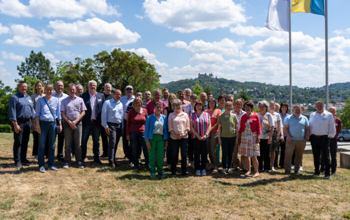 Eine bunt gemischte Gruppe von Menschen steht unter blauem Himmel auf einer Wiese im Freien zusammen und verkörpert den Geist der Hochschule Coburg. Sie posieren vor einer malerischen Kulisse mit Bäumen und einem Hügel in der Ferne, während auf der rechten Seite des Bildes mehrere Flaggen stolz wehen.