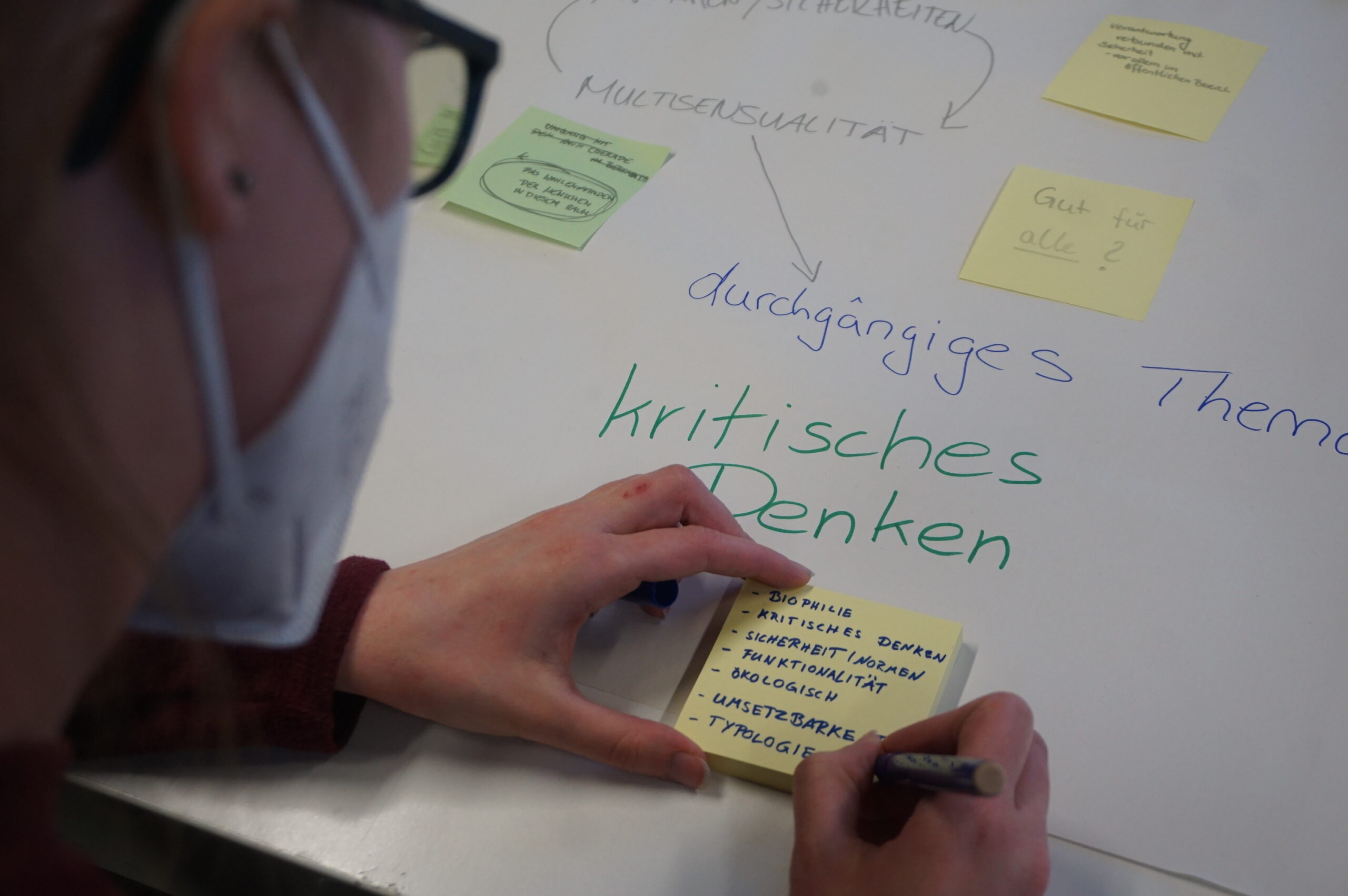 Eine Person mit Brille und Maske schreibt auf einen Haftzettel an einem Tisch, der mit verschiedenen Haftzetteln geschmückt ist. Unter ihnen sind die deutschen Worte „kritisches Denken“ zu sehen, was den Schwerpunkt auf kritisches Denken widerspiegelt, der an der Hochschule Coburg betont wird.
