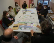 Eine Gruppe von Menschen, einige davon mit Masken, sitzt in der Hochschule Coburg um einen mit Papieren und Diagrammen bedeckten Tisch. In dem hell erleuchteten Raum scheinen sie an einer Diskussion oder Besprechung teilzunehmen.