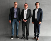 Drei Männer stehen in einer Reihe vor einer grauen Wand. Sie tragen Business-Kleidung mit Anzügen und Elementen im Smart-Casual-Stil. Das Bild lässt auf eine formelle Zusammenkunft schließen, möglicherweise auf einer Veranstaltung der Hochschule Coburg.