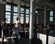 In einem Raum mit großen Fenstern der Hochschule Coburg sitzen Menschen, einige unterhalten sich. Zwei Personen stehen vorne und unterhalten sich. Der Raum verfügt über eine Bühne und eine moderne Beleuchtung. Sonnenlicht fällt durch die Fenster und sorgt für eine helle Atmosphäre.