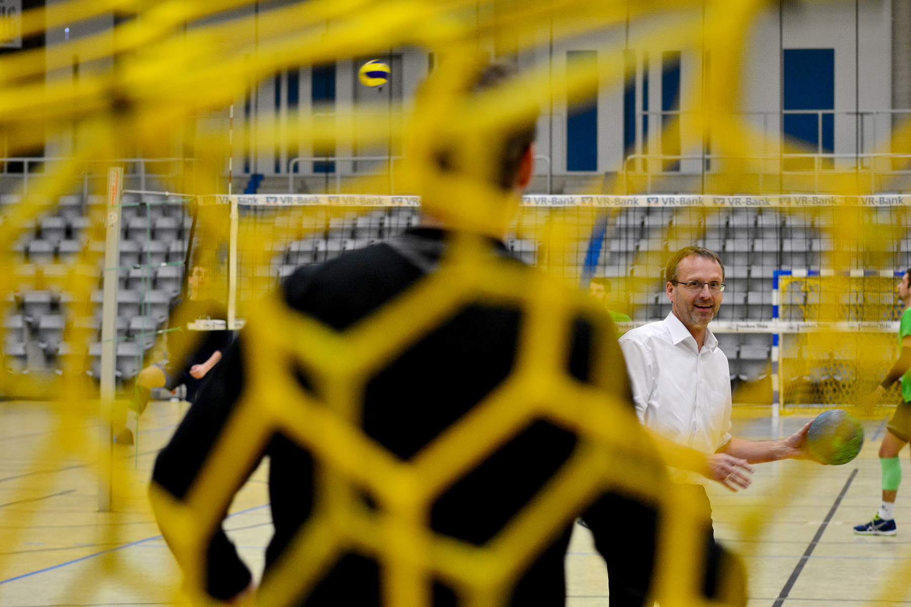 Ein Mann in weißem Hemd steht lächelnd auf dem Sportplatz der Hochschule Coburg und hält einen Fußball in der Hand, der durch ein gelbes Netz zu sehen ist. Eine andere Person, leicht verschwommen, steht ihm gegenüber. Im Hintergrund sind leere Zuschauerränge und ein Volleyball in der Luft zu sehen.