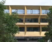 Die Hochschule Coburg verfügt über ein modernes Gebäude mit einer einzigartigen goldenen Fassade und großen kubischen Rahmen um die Fenster, die teilweise von üppigen grünen Bäumen im Vordergrund verdeckt werden, wodurch eine harmonische Verbindung aus Architektur und Natur entsteht.
