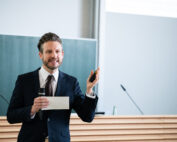 Ein Mann im Anzug hält in einem Hörsaal der Hochschule Coburg einen Vortrag. Er hält Mikrofon und Zeigegerät in der Hand und steht selbstbewusst vor der Tafel. Der Raum strahlt eine helle und professionelle Atmosphäre aus, perfekt für akademische Erkundungen.