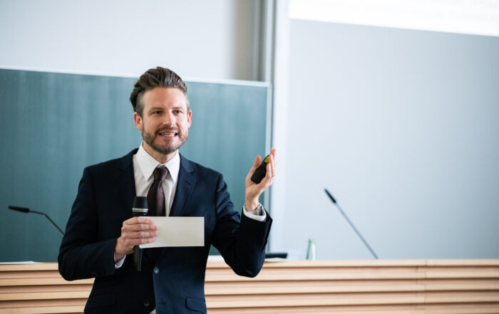 Ein Mann im Anzug hält in einem Hörsaal der Hochschule Coburg einen Vortrag. Er hält Mikrofon und Zeigegerät in der Hand und steht selbstbewusst vor der Tafel. Der Raum strahlt eine helle und professionelle Atmosphäre aus, perfekt für akademische Erkundungen.