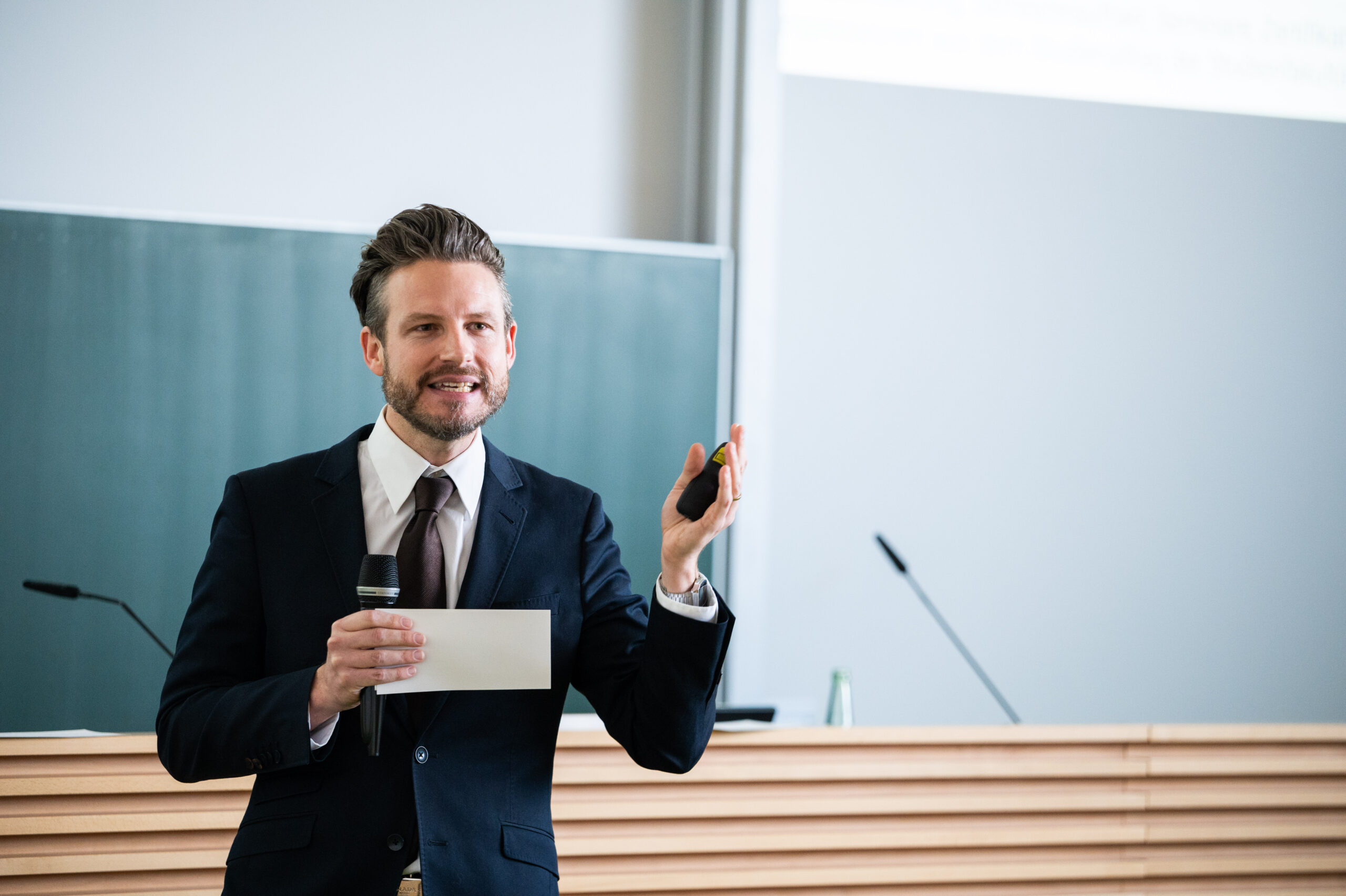 Ein Mann im Anzug hält in einem Hörsaal der Hochschule Coburg einen Vortrag. Er hält Mikrofon und Zeigegerät in der Hand und steht selbstbewusst vor der Tafel. Der Raum strahlt eine helle und professionelle Atmosphäre aus, perfekt für akademische Erkundungen.
